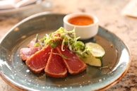 Several slices of seared tuna are artfully arranged on a ceramic plate. The tuna is garnished with fresh microgreens, and there are lemon or lime wedges on the side. A small white ramekin containing a bright orange dipping sauce is also present on the plate. The background is a textured countertop with muted colors.