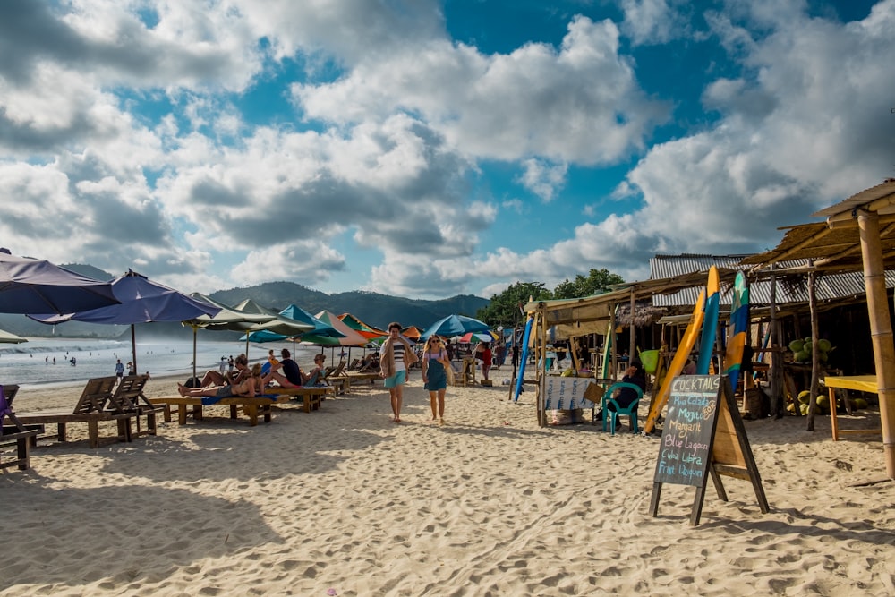 Dos personas caminando por la playa con sombrillas variadas y cabañas de nipa