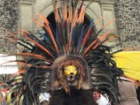 An elaborate ceremonial headdress adorned with large colorful feathers in shades of orange, black, and blue. The centerpiece features a jaguar face with vivid yellow and black markings, positioned in front of a textured stone building.