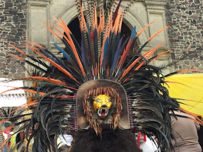An elaborate ceremonial headdress adorned with large colorful feathers in shades of orange, black, and blue. The centerpiece features a jaguar face with vivid yellow and black markings, positioned in front of a textured stone building.