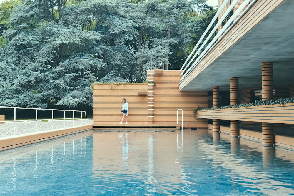 흰 셔츠를 입은 여자가 낮에는 갈색 벽돌 벽과 나무 근처의 푸른 물 수영장 옆에 서 있습니다.