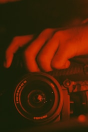 A close-up view of a camera lens with a hand gripping the camera body. The image is dominated by a reddish-orange hue, creating a warm and intense atmosphere.