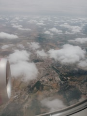 An aerial view of a landscape partially obscured by clouds, with a visible airplane wing and engine. The ground below includes sprawling fields, a mix of farmland and urban areas.