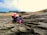 A group of children crouching and exploring the rocky terrain by the coast. They appear curious, examining the natural features and tidal pools. The landscape is rugged with visible rock formations, and the sky is partially cloudy with some clear visibility of distant trees and mountains.