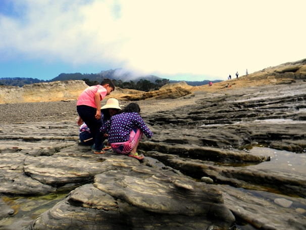 A group of children crouching and exploring the rocky terrain by the coast. They appear curious, examining the natural features and tidal pools. The landscape is rugged with visible rock formations, and the sky is partially cloudy with some clear visibility of distant trees and mountains.