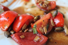 A close-up view of sliced tomatoes topped with various seeds, drizzled with a rich brown sauce. The tomatoes are vibrant red, and the seeds vary in color, some appearing dark and others light with a glossy finish. The dish seems fresh and appetizing.