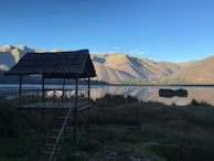 A rustic wooden structure with a thatched roof is situated by a tranquil lake. The water reflects the surrounding mountains and clear blue sky. Grassy vegetation surrounds the area, creating a natural and serene environment.