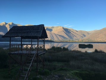 A rustic wooden structure with a thatched roof is situated by a tranquil lake. The water reflects the surrounding mountains and clear blue sky. Grassy vegetation surrounds the area, creating a natural and serene environment.