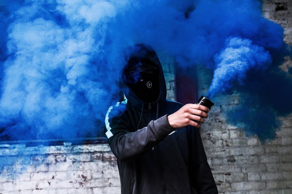 persona coperta con maschera e felpa con cappuccio che usa il fumo blu