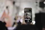 A hand holding a smartphone captures a scene with people on stage, possibly a performance or event. The background is blurred, focusing on the phone's screen, which shows a clearer image of the event.