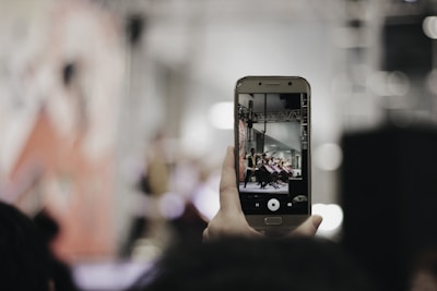 A hand holding a smartphone captures a scene with people on stage, possibly a performance or event. The background is blurred, focusing on the phone's screen, which shows a clearer image of the event.
