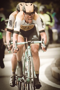 Two cyclists are in motion, wearing matching beige helmets and jerseys with logos. The cyclist in front is visibly exerting effort, gripping the handlebars tightly and has an intense expression. They are in a competitive race setting with blurred figures and greenery in the background.
