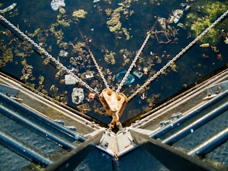 A body of water is filled with scattered debris and green algae. Chains extend from a metallic structure, converging at a buoy or anchor point. The water surface reflects sunlight, highlighting various pieces of floating litter.
