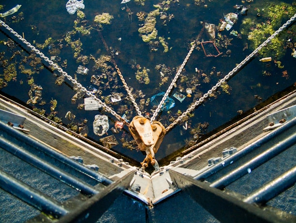 A body of water is filled with scattered debris and green algae. Chains extend from a metallic structure, converging at a buoy or anchor point. The water surface reflects sunlight, highlighting various pieces of floating litter.