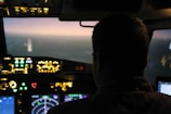 A person is sitting in an aircraft cockpit, focusing on the illuminated control panels and screens. The view through the cockpit window shows a horizon, hinting at either dawn or dusk lighting.