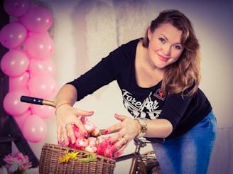 A woman with long, light brown hair is leaning slightly forward, smiling warmly while arranging a bouquet of pink roses in a wicker basket attached to a bicycle. Pink balloons are arranged in an arch on the left side, adding a festive atmosphere. She is wearing a black long-sleeve shirt and blue jeans, adorned with gold bracelets and rings.