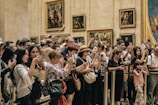A large group of people stands closely together, many holding up cameras and smartphones to take pictures. They appear to be in an art gallery, with several framed paintings visible on the walls behind them. The crowd seems diverse, including both adults and children, capturing a moment of collective interest.