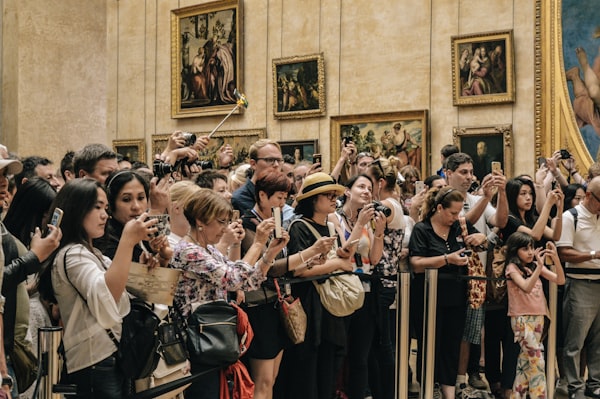 a crowd of people in an art gallery hold up phones and cameras towards a work off to the right