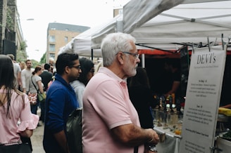 A crowd of people gather at an outdoor market under white tents, which appear to be food stalls. An older man with white hair and glasses, wearing a pink polo shirt, is reading a menu at a stall. There are various people in casual clothing browsing the market, and the background shows a mix of urban buildings.