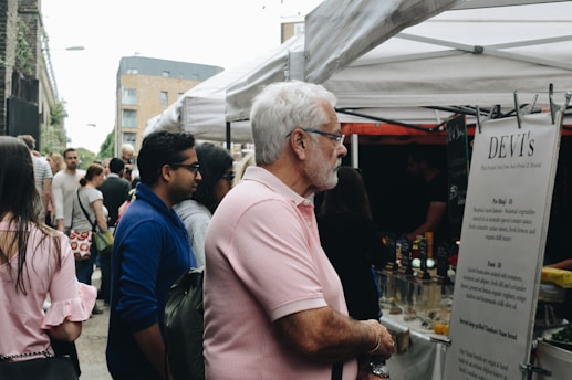 A crowd of people gather at an outdoor market under white tents, which appear to be food stalls. An older man with white hair and glasses, wearing a pink polo shirt, is reading a menu at a stall. There are various people in casual clothing browsing the market, and the background shows a mix of urban buildings.