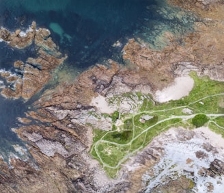 An aerial view of a coastal area featuring rocky outcrops and green vegetation. The land is intersected by winding paths, leading to a small building near the shore. The shoreline consists of a mix of rocks and sandy patches, with the ocean displaying shades of blue and turquoise.