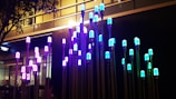 A series of illuminated light fixtures with colorful bulbs are mounted on tall poles of varying heights. The colors of the lights range from blue to purple, casting a vibrant glow. The fixtures are installed against an urban backdrop with a commercial building and trees partially visible.