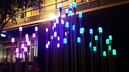 A series of illuminated light fixtures with colorful bulbs are mounted on tall poles of varying heights. The colors of the lights range from blue to purple, casting a vibrant glow. The fixtures are installed against an urban backdrop with a commercial building and trees partially visible.