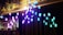 A series of illuminated light fixtures with colorful bulbs are mounted on tall poles of varying heights. The colors of the lights range from blue to purple, casting a vibrant glow. The fixtures are installed against an urban backdrop with a commercial building and trees partially visible.