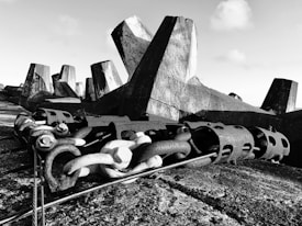A black and white image featuring large concrete structures with sharp, angular shapes. Metal chains and industrial connectors are visible in the foreground, laid on a textured surface. The background shows an arrangement of similar concrete forms under a cloudy sky.