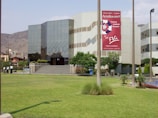 A modern building with reflective glass windows and geometric patterns, surrounded by a well-maintained grassy lawn and a few trees. A large sign with red and white colors is prominently displayed on the building's facade. Several people are standing near a pathway.