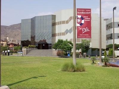 A modern building with reflective glass windows and geometric patterns, surrounded by a well-maintained grassy lawn and a few trees. A large sign with red and white colors is prominently displayed on the building's facade. Several people are standing near a pathway.