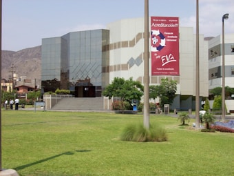 A modern building with reflective glass windows and geometric patterns, surrounded by a well-maintained grassy lawn and a few trees. A large sign with red and white colors is prominently displayed on the building's facade. Several people are standing near a pathway.
