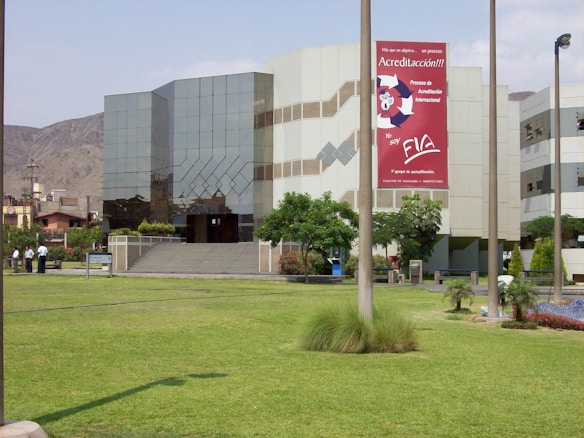 A modern building with reflective glass windows and geometric patterns, surrounded by a well-maintained grassy lawn and a few trees. A large sign with red and white colors is prominently displayed on the building's facade. Several people are standing near a pathway.