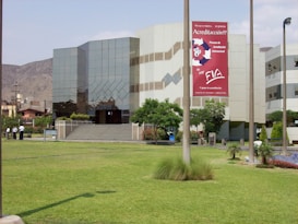 A modern building with reflective glass windows and geometric patterns, surrounded by a well-maintained grassy lawn and a few trees. A large sign with red and white colors is prominently displayed on the building's facade. Several people are standing near a pathway.