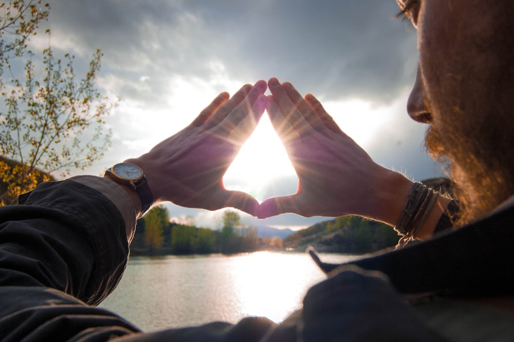 A bearded man beside a lake holds his hands in a triangle, the sun shining through