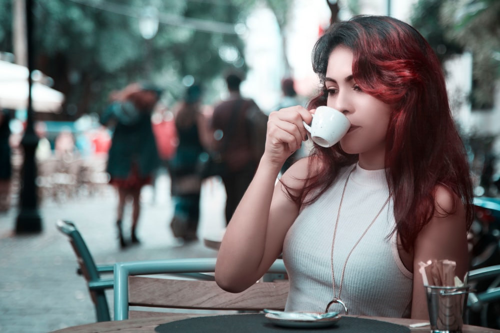 A woman with dyed red hair sips from a teacup at an outdoor cafe table