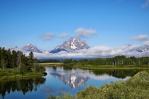 Majestic mountains rise in the background with their snow-capped peaks partially obscured by light clouds. A pristine lake reflects the scenery, bordered by lush green forests and vegetation. Two small kayaks are visible on the lake, adding a touch of activity to the tranquil setting.