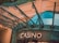 The image displays the entrance to a casino, characterized by detailed and ornate architectural elements with green metal structures and a sign that reads 'CASINO'. The background includes a glass dome and a peach-colored wall.