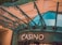 The image displays the entrance to a casino, characterized by detailed and ornate architectural elements with green metal structures and a sign that reads 'CASINO'. The background includes a glass dome and a peach-colored wall.