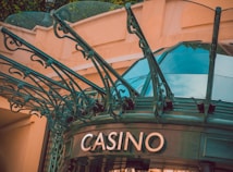 The image displays the entrance to a casino, characterized by detailed and ornate architectural elements with green metal structures and a sign that reads 'CASINO'. The background includes a glass dome and a peach-colored wall.