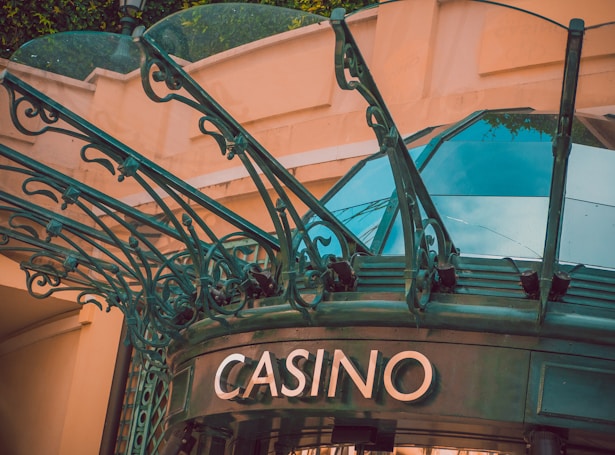 The image displays the entrance to a casino, characterized by detailed and ornate architectural elements with green metal structures and a sign that reads 'CASINO'. The background includes a glass dome and a peach-colored wall.