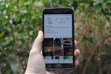 A hand is holding a Samsung smartphone displaying an Instagram profile with various posts and details visible on the screen. The background consists of blurred greenery and foliage, suggesting an outdoor setting.