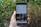 A hand is holding a Samsung smartphone displaying an Instagram profile with various posts and details visible on the screen. The background consists of blurred greenery and foliage, suggesting an outdoor setting.