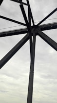 Steel beams form a geometric pattern against a cloudy, overcast sky. The structure appears industrial, suggesting it may be part of a bridge or tower.