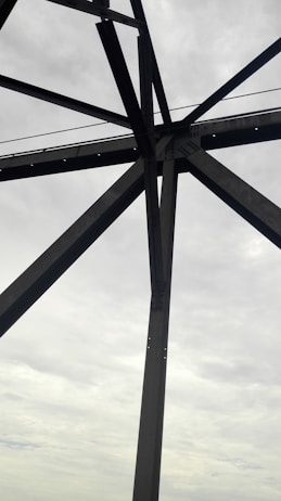Steel beams form a geometric pattern against a cloudy, overcast sky. The structure appears industrial, suggesting it may be part of a bridge or tower.
