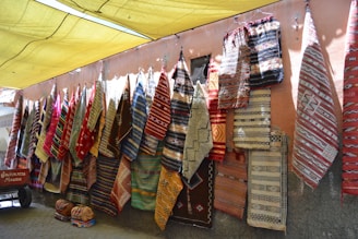 A colorful display of various woven rugs hanging on a wall, featuring intricate patterns and a variety of colors. Some rugs have geometric designs, while others include more intricate, detailed motifs. The scene is set in an outdoor marketplace with a yellow canopy above providing shade.