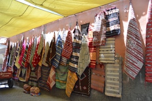 A colorful display of various woven rugs hanging on a wall, featuring intricate patterns and a variety of colors. Some rugs have geometric designs, while others include more intricate, detailed motifs. The scene is set in an outdoor marketplace with a yellow canopy above providing shade.