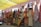 A colorful display of various woven rugs hanging on a wall, featuring intricate patterns and a variety of colors. Some rugs have geometric designs, while others include more intricate, detailed motifs. The scene is set in an outdoor marketplace with a yellow canopy above providing shade.
