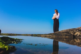 A woman stands on a rocky outcrop next to a serene body of water, cradling her pregnant belly. The sky is clear and blue, and her reflection is visible in the water. She wears a white blouse and a long black skirt, conveying a sense of calm and introspection.