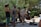 A group of street musicians are performing outdoors surrounded by greenery. The performers include a trombone player wearing a sun hat, a saxophonist, an upright bass player, and a guitarist who appears lively and animated. Equipment cases and a small box for donations are on the ground nearby.